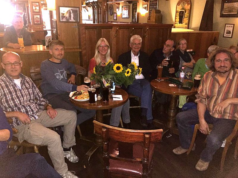 Bild von mehreren Personen, die um einen Tisch im Pub sitzen. Auf dem Tisch stehen Sonnenblumen.