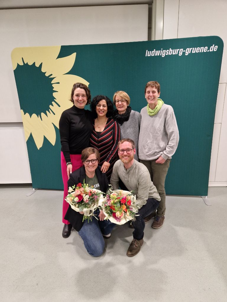 Gruppenfoto vor einer Fotowand der Grünen; zwei Personen halten Blumensträuße in der Hand.