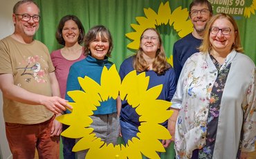 Das Bild zeigt sechs Personen mit einer großen hölzernen Sonnenblume vor einem grünen Hintergrund.