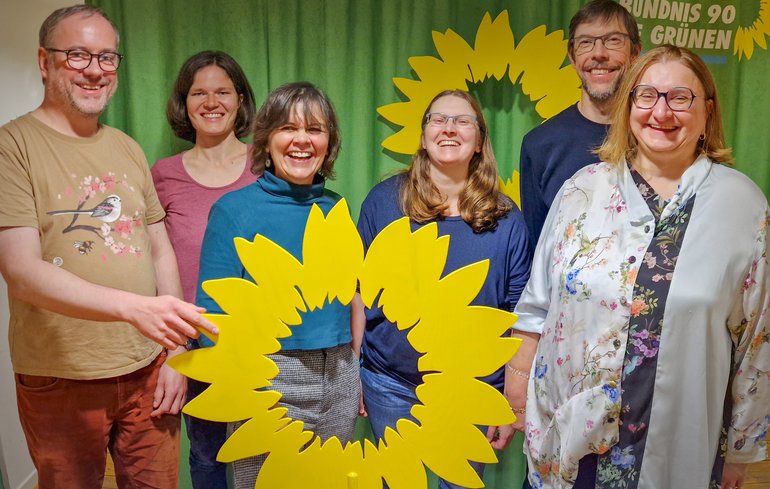 Das Bild zeigt sechs Personen mit einer großen hölzernen Sonnenblume vor einem grünen Hintergrund.