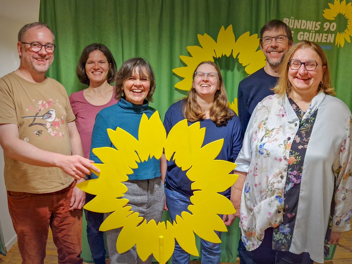 Das Bild zeigt sechs Personen mit einer großen hölzernen Sonnenblume vor einem grünen Hintergrund.