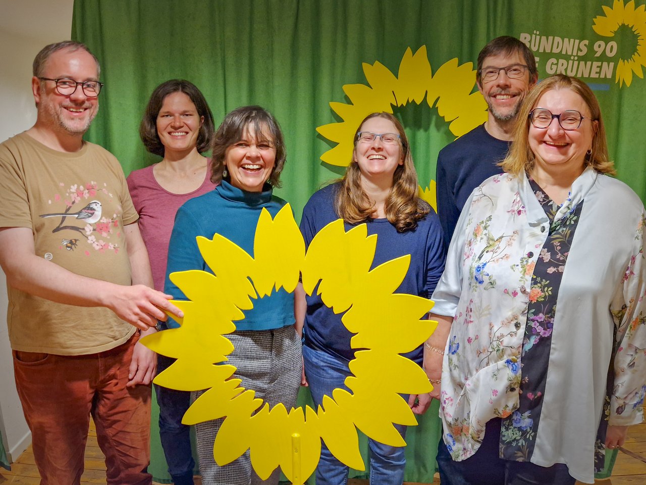 Das Bild zeigt sechs Personen mit einer großen hölzernen Sonnenblume vor einem grünen Hintergrund.