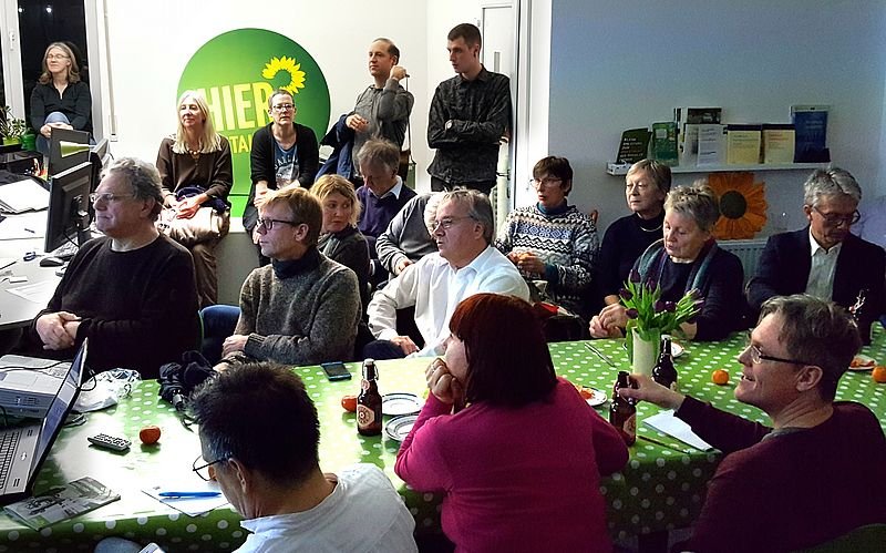 Ca. 20 Menschen sitzen und stehen in einem Raum. Auf den Tischen in der Mitte liegt eine grüne Tischdecke.