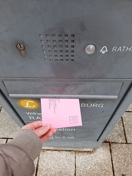 Bild von einer Hand, die einen rosafarbenen Umschlag in einen Briefkasten mit dem Logo der Stadt Ludwigsburg einwirft.