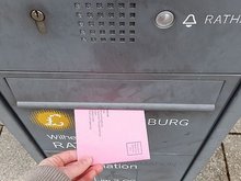 Bild von einer Hand, die einen rosafarbenen Umschlag in einen Briefkasten mit dem Logo der Stadt Ludwigsburg einwirft.