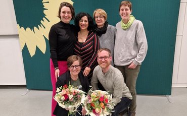 Gruppenfoto vor einer Fotowand der Grünen; zwei Personen halten Blumensträuße in der Hand.